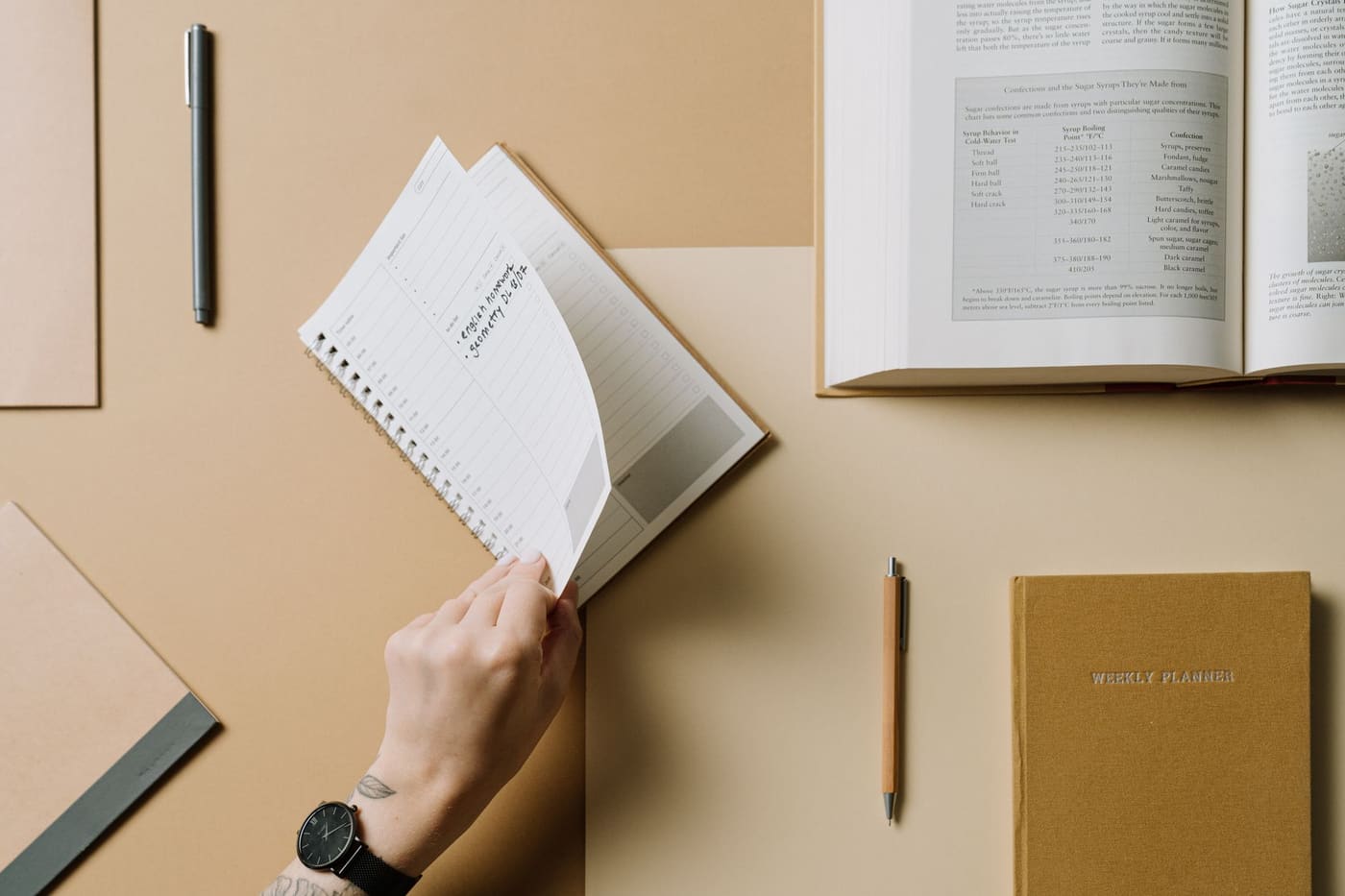 A hand turning a notebook page beside a planner and an open book
