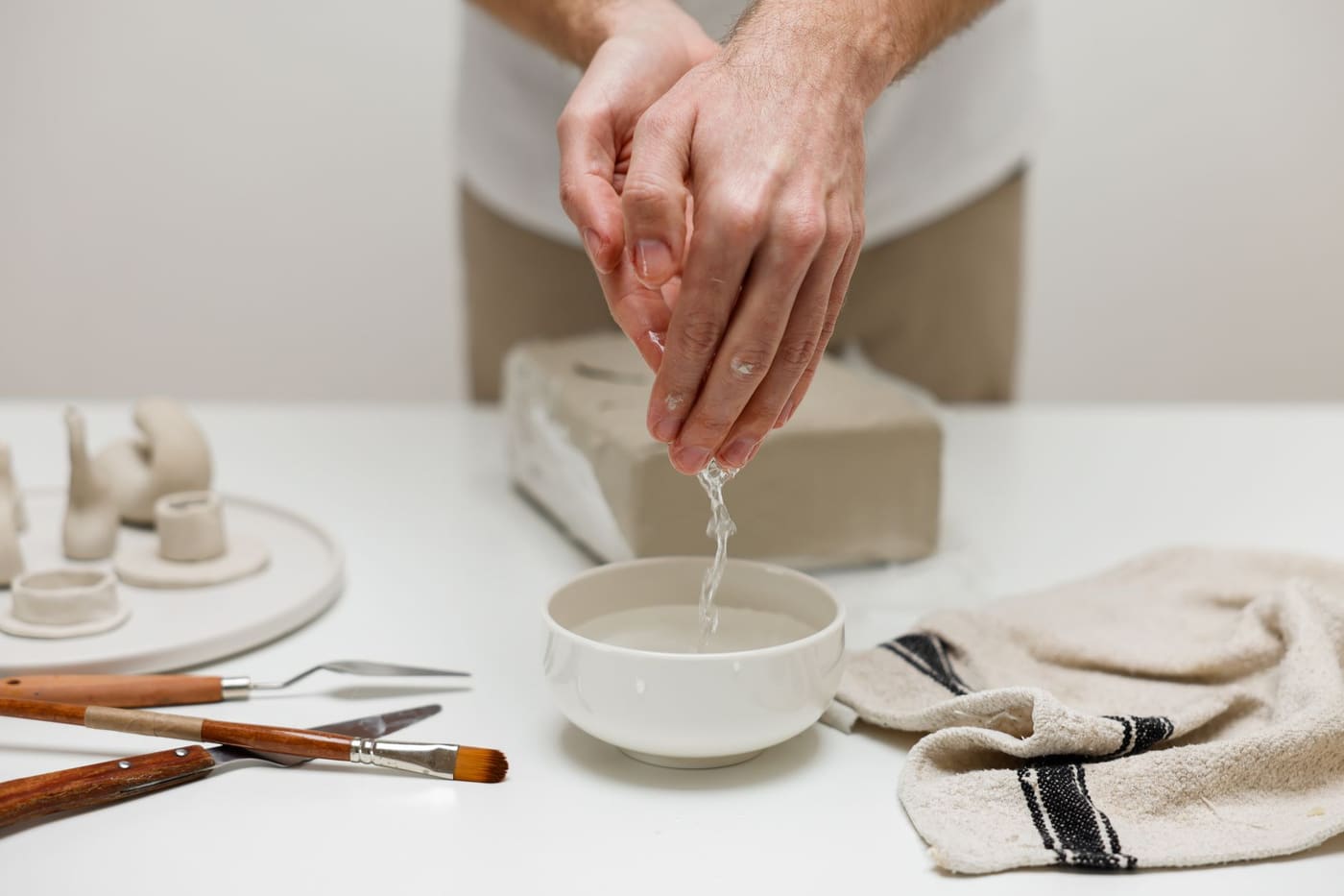 Hands crafting ceramics with tools, a bowl of water and a block of clay