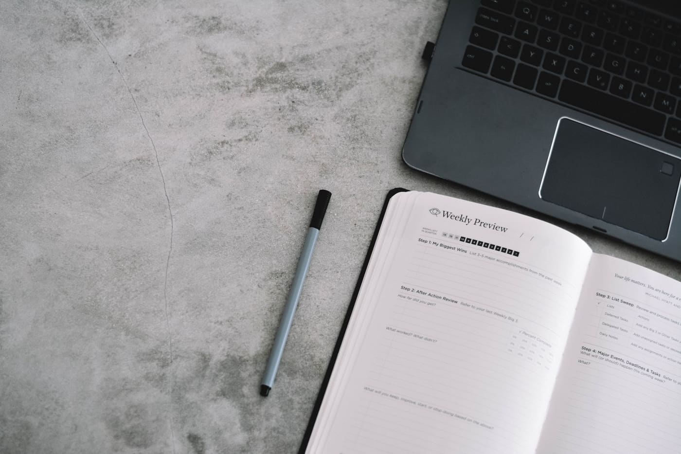 A minimalist desk with a planner, pen and laptop on a gray surface