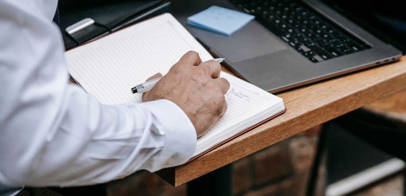 A hand writing in a notebook beside a laptop