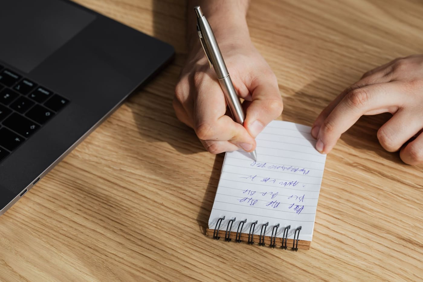 A hand writing in a small spiral notebook next to a laptop on a wooden table