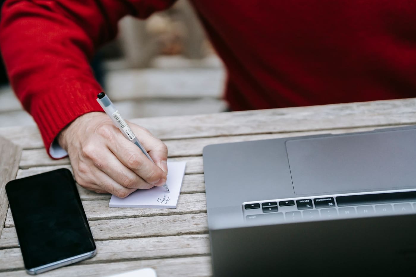 A hand writing a note on paper beside a laptop and a phone on a wooden table