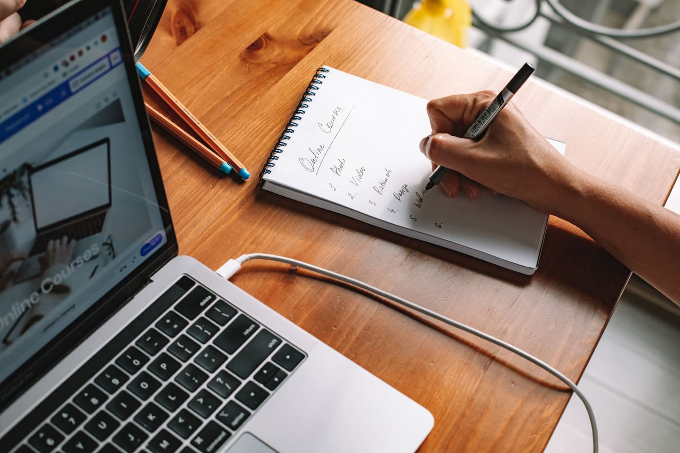 A person writing in a notebook next to a laptop at a wooden desk